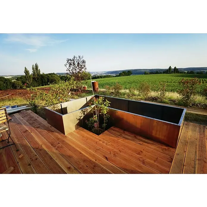 Palatino Hochbeet Urban L - 300 x 200 x 70 cm, Cortenstahl, Rostbraun - Eine erhöhte Pflanzkiste aus Cortenstahl sitzt auf einer Holzterrasse mit Blick auf eine grüne Landschaft. Der Pflanzkasten enthält Pflanzen.