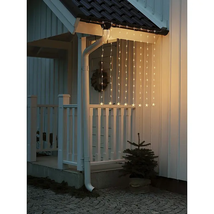 Konstsmide LED-Lichtervorhang - Außen, 105 cm, Bernstein, 96 Stk., Batteriebetrieben - Sternen-Lichterketten schmücken die Veranda und sorgen für eine festliche Atmosphäre. Die warmen Lichter leuchten hell vor dem weißen Haus.