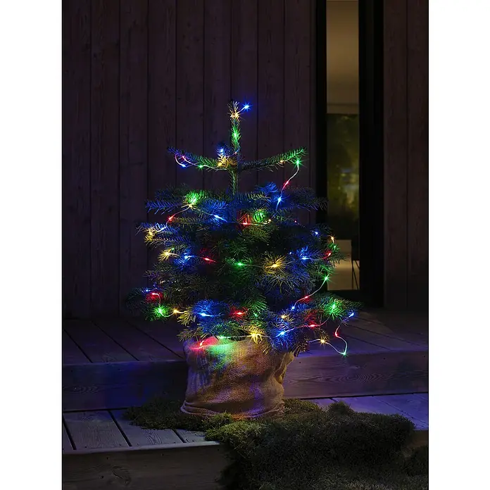 Konstsmide LED-Lichterkette - Farblos, Außen, Lichtfarbe: Mehrfarbig, Anzahl Leuchtmittel: 80 Stk., Batteriebetrieben - Stimmungsvolle Aufnahme eines kleinen Weihnachtsbaums mit bunten Lichtern auf einer Holzterrasse, perfekt für festliche Dekoration.