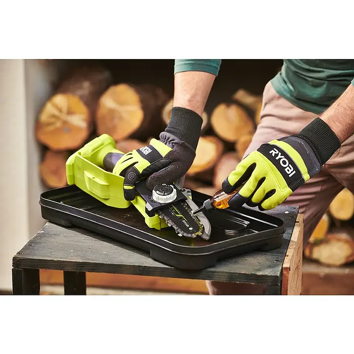 Ryobi ONE+ Akku-Astsäge RY18PSX10A-120 - 18 V, 2 Ah, Li-Ionen, 1 Akku, Schwertlänge: 10 cm - Eine Person, die eine tragbare Ryobi-Kettensäge ölt. Die Säge steht auf einem Tisch vor einem Stapel Holzscheite.