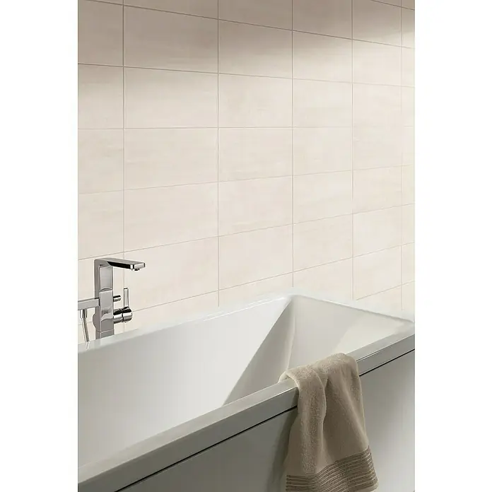 Wandfliese Leyre Arena Beige - 30 x 60 cm, Beige, Glänzend - Nahaufnahme einer Badewanne mit Armatur und Handtuch vor einer gefliesten Wand.