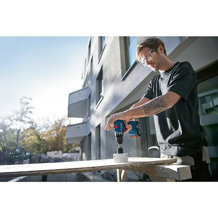 Bosch Professional AMPShare 18V Akku-Bohrschrauber GSR 18V-45 - 18 V, Ohne Akku, 45 Nm, Ohne L-Boxx 136 - Ein Mann bohrt mit einer blauen Bosch Bohrmaschine ein Loch in ein Holzbrett, das auf einer Werkbank liegt, während er eine Schutzbrille trägt.
