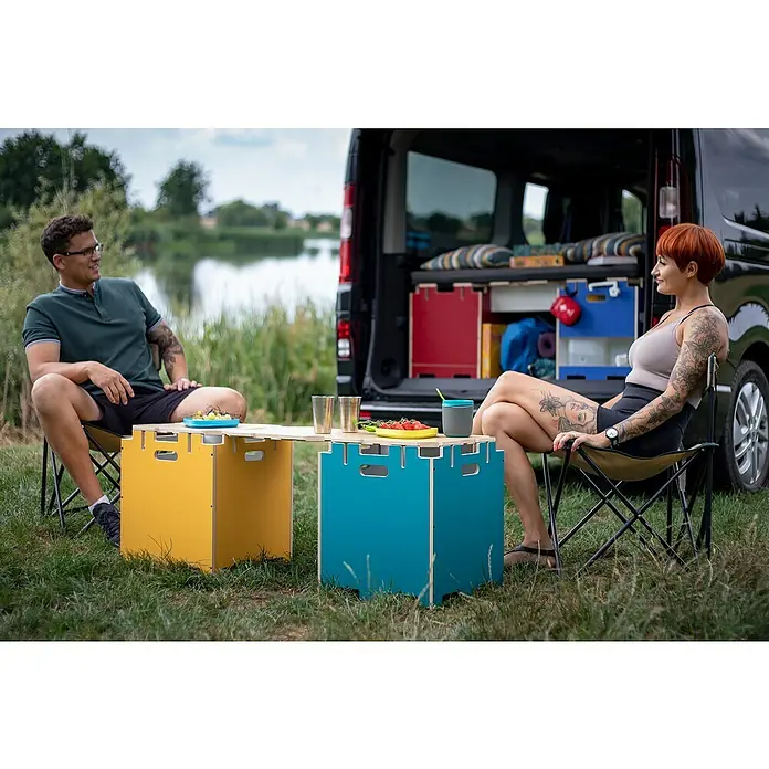 Vojo Camper Ausbau-Set Base L - 13 -tlg., Geeignet für: Transporter - Zwei Personen sitzen an einem farbenfrohen Campingtisch und genießen die Natur neben einem schwarzen Van.