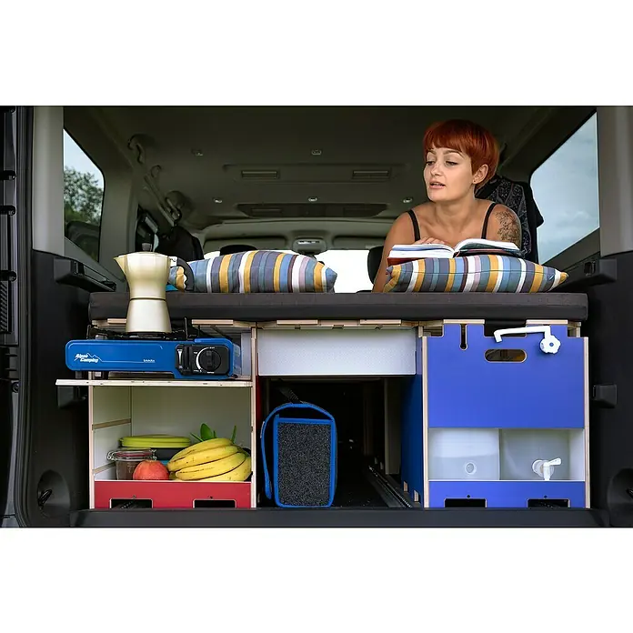 Vojo Camper Ausbau-Set Nomad - 23 -tlg., Geeignet für: Transporter - Frau liest in einem umgebauten Van mit Küchenzeile, Kocher, Wasserkanister und Bett für Campingausflüge.