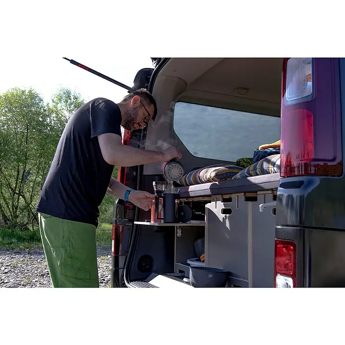 Vojo Camper Ausbau-Set Base L - 13 -tlg., Geeignet für: Transporter - Ein Mann gießt Wasser aus einem Wasserkocher in eine Tasse in einem umgebauten Van, mit eingebauter Küche und Bett.
