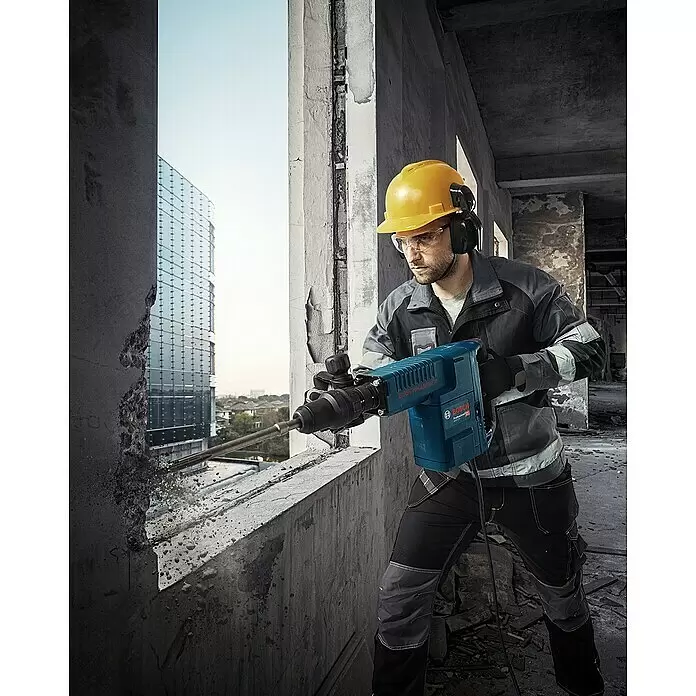 Bosch Professional Martillo demoledor GSH11E - 1.500 W, 16,8 J, Número máx. de impactos: 1.890 r.p.m. - Hombre con equipo de construcción usa un martillo perforador Bosch azul para romper hormigón de un muro, revelando una vista urbana.