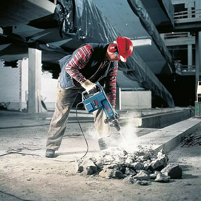 Bosch Professional Martillo demoledor GSH11E - 1.500 W, 16,8 J, Número máx. de impactos: 1.890 r.p.m. - Obrero con equipo de protección utiliza un martillo percutor Bosch azul para romper hormigón. El polvo se eleva.