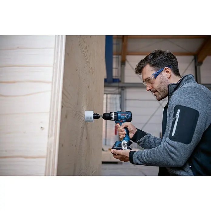 Bosch Professional AMPShare 18V Taladro atornillador percutor de batería GSB18V-90 - 18 V, 2 baterías, 4 Ah, 64 Nm, Incl. L-Boxx 136 + 2 baterías recargables 18 V/4 Ah - Un artesano con gafas protectoras está perforando un agujero en una pared de madera con un taladro inalámbrico azul Bosch Professional.