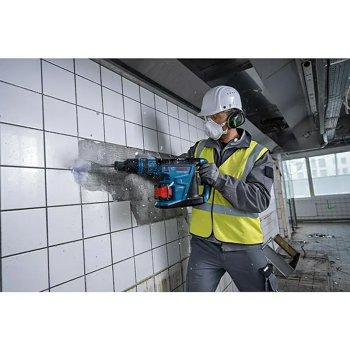 Bosch Professional AMPShare 18V Akku-Kombihammer GBH 18V-40 C - 18 V, 2 Akkus, 8 Ah, 9 J - Ein Bauarbeiter mit Schutzhelm und Maske benutzt einen Bosch-Bohrhammer, um Fliesen von einer Wand zu entfernen. Die Bosch-Maschine ist blau und schwarz.