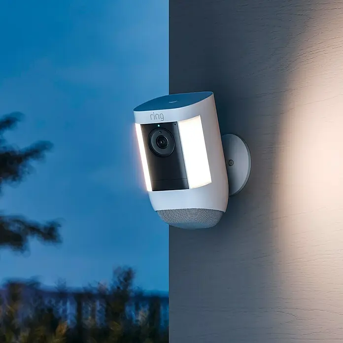 Ring Überwachungskamera Spotlight Cam Pro Battery - Weiß, Auflösung: 1.080 Pixel (Full HD), Akku - Nahaufnahme einer Ring Spotlight Cam, die an einer Wand montiert ist und nachts leuchtet, für erhöhte Sicherheit.\n