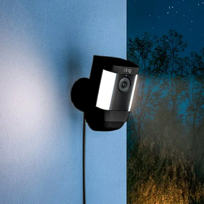 Ring Überwachungskamera Spotlight Cam Pro Plugin - Schwarz, Auflösung: 1.080 Pixel (Full HD), Netzanschluss - Nahaufnahme einer Ring-Überwachungskamera mit eingeschaltetem Flutlicht, montiert an einer Wand, mit Blick auf einen nächtlichen Außenbereich.