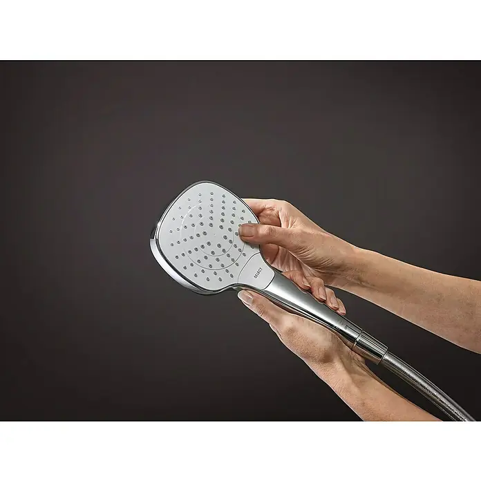 Hansgrohe Croma Select E Brausegarnitur 110 Vario - Lochabstand: 62,5 cm, Art Strahl: Rain, TurboRain, IntenseRain, Chrom/Weiß, Wassersparfunktion - Eine Handbrause mit Chromfinish wird von zwei Händen vor einem dunklen Hintergrund gehalten. Die Brause verfügt über verschiedene Strahlarten.