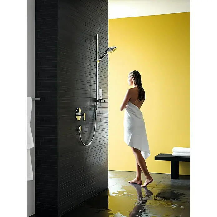 Hansgrohe Raindance Select S Brausegarnitur 150 - Lochabstand: 62,5 cm, Art Strahl: RainAir, CaresseAir, Mix, Chrom, Seifenschale - Eine Frau steht in einem Badezimmer mit einer modernen Duscharmatur mit goldenen Elementen und schwarzen Fliesen.