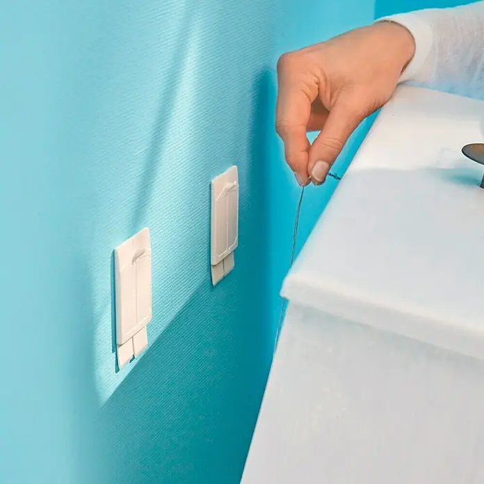 Tesa Clavo adhesivo - Blanco, Carga soportada: 2 kg, 2 ud., Paredes pintadas - Una mano sujeta un cuadro a una pared azul claro con ganchos adhesivos. Los ganchos son discretos y fáciles de instalar.