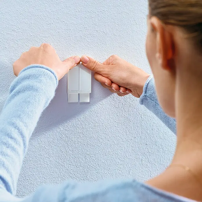 Tesa Clavo adhesivo - Blanco, Carga soportada: 2 kg, 2 ud., Paredes pintadas - Una mujer fija un gancho adhesivo blanco y rectangular a una pared azul claro para colgar objetos.