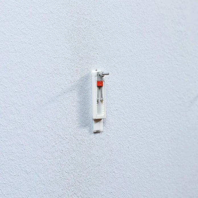 Tesa Tornillo adhesivo ajustable - Rectangular, Carga soportada: 1 kg, 2 ud. - Primer plano de un colgador de cuadros blanco fijado a la pared, con un botón rojo y un gancho gris.