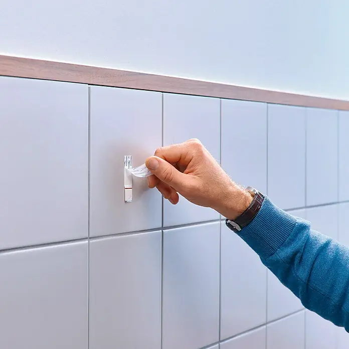 Tesa Clavo adhesivo - Blanco, Carga soportada: 2 kg, 2 ud., Azulejos - Una mano retira la película protectora de un gancho adhesivo, montado en una pared alicatada. Fácil montaje para accesorios de baño.