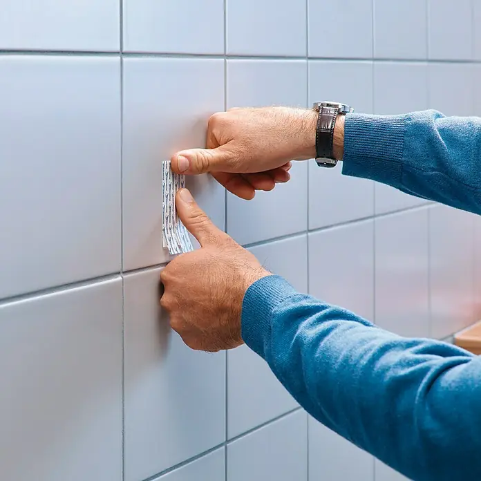 Tesa Clavo adhesivo - Blanco, Carga soportada: 4 kg, 2 ud. - Un hombre adhiere una tira adhesiva a una pared de azulejos, mostrando una manera fácil de colgar objetos sin dañar las superficies.