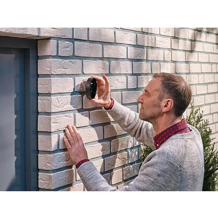 Tesa Tornillo adhesivo - Triangular, Carga soportada: 5 kg, 2 ud. - Un hombre sujeta un soporte de montaje negro a una pared de ladrillo al lado de una puerta para instalar un dispositivo electrónico.