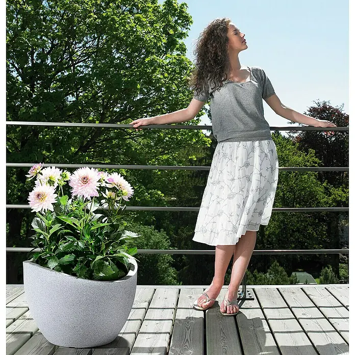 Scheurich Wave Globe Pflanztopf - Außenmaß (Ø x H): 39,5 x 29,3 cm, Kunststoff, Steingrau - Das Bild zeigt einen stilvollen grauen Pflanztopf mit rosa Dahlien, der auf einer Holzterrasse neben einer Frau steht.