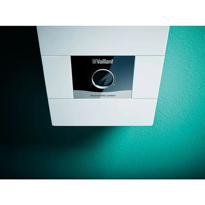 Vaillant Durchlauferhitzer VED E 21/8C COMFORT - 21 kW, Elektronisch, Geschlossen - Nahaufnahme einer Vaillant electronicVED comfort Warmwasserheizung mit schwarzer Blende und Bedienknopf, montiert an einer farbigen Wand.