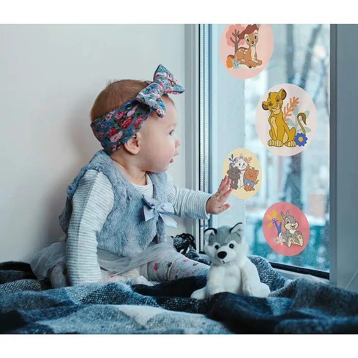 Komar Wandtattoo Disney Better Together Bubbles30 x 30 cm - Ein bezauberndes Baby mit einem blumigen Stirnband sitzt auf einem Fensterbrett und betrachtet Fensteraufkleber mit Disney-Figuren.