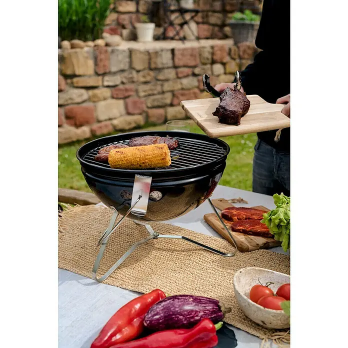 Kingstone Kugelgrill Cutie Pie - Durchmesser Grillfläche: 37 cm, Schwarz - Ein Holzkohlegrill mit Grillgut wie Mais und Steaks wird gezeigt, während eine Person mit einer Zange ein Steak auf ein Schneidebrett legt.