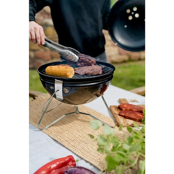 Kingstone Kugelgrill Cutie Pie - Durchmesser Grillfläche: 37 cm, Schwarz - Ein tragbarer Grill mit Maiskolben und Fleisch, eine Person hält den Deckel und die Zange. Köstliches Grillgut für den Sommer.