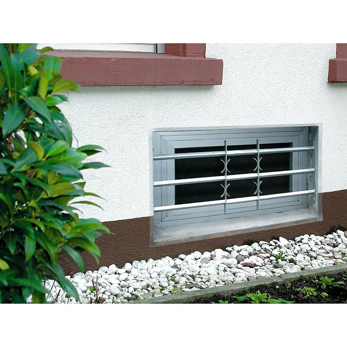 Stabilit Fenstergitter - Höhe: 450 mm, Auszugslänge: 50 cm - 65 cm, Fensterlaibungsmontage - Nahaufnahme eines Kellerfensters mit Metallgitter zum Schutz vor Einbruch und zur Sicherheit. Steine umranden das Fenster.