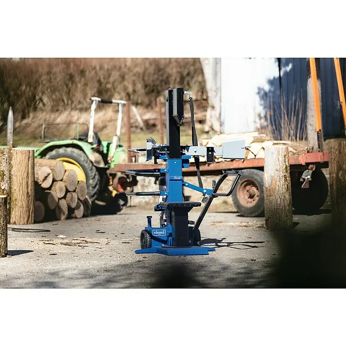 Scheppach Holzspalter Compact 10t 400V - 3.500 W, 10 t, Max. Spaltgutlänge: 106 cm - 750 mm - Nahaufnahme eines blauen Scheppach Holzspalters vor einem Schlepper und einem Holzanhänger im Außenbereich.