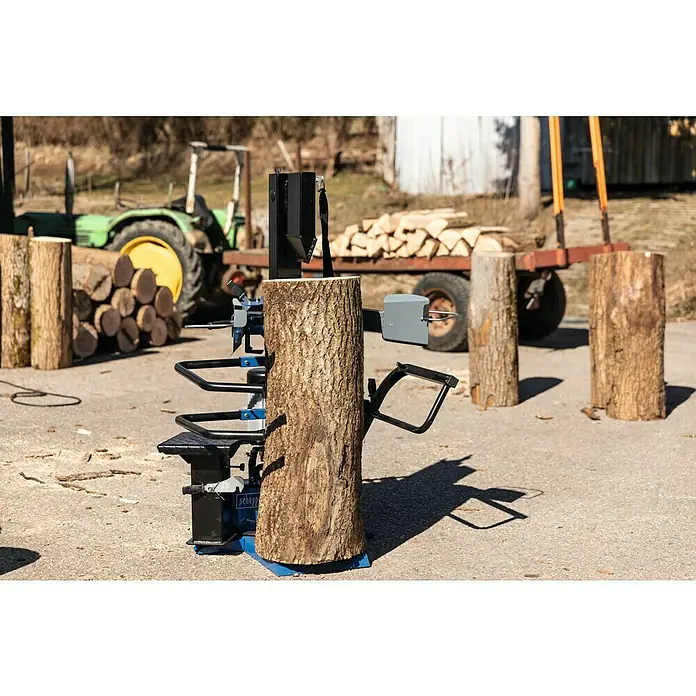 Scheppach Holzspalter Compact 10t 400V - 3.500 W, 10 t, Max. Spaltgutlänge: 106 cm - 750 mm - Ein Scheppach Holzspalter steht bereit, um einen dicken Baumstamm zu bearbeiten, während im Hintergrund ein Traktor und Holzstapel zu sehen sind.