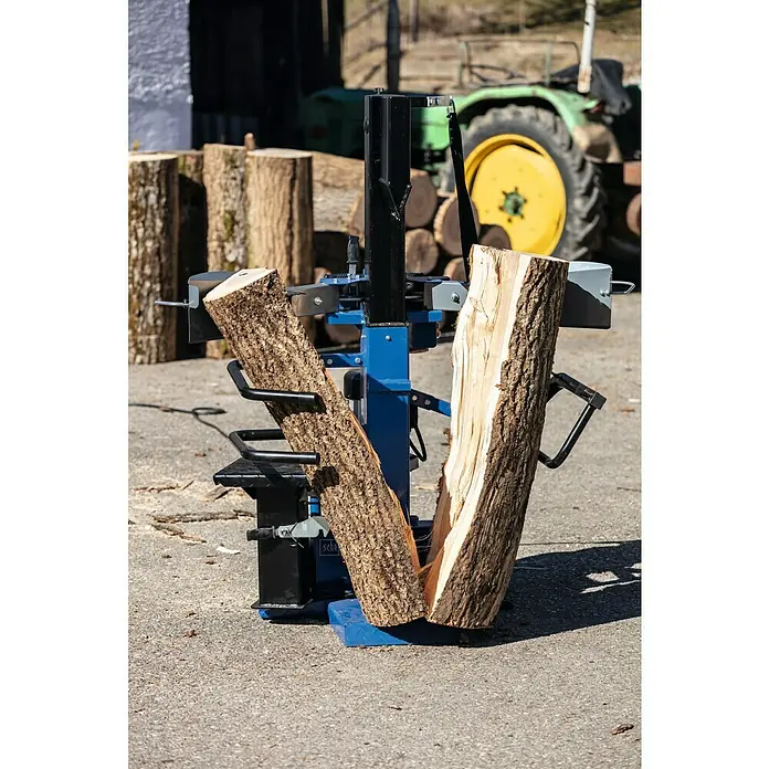 Scheppach Holzspalter Compact 10t 400V - 3.500 W, 10 t, Max. Spaltgutlänge: 106 cm - 750 mm - Ein Scheppach Holzspalter in Aktion, der ein Holzstück spaltet, mit Holzstapeln und einem Traktor im Hintergrund.