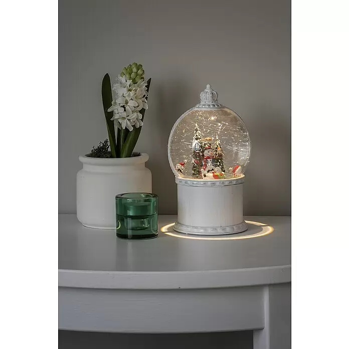 Konstsmide LED-Schneelaterne Schneemann mit Baum - Innen, Altweiß, 12,5 x 12,5 x 21 cm, Schneemann mit Christbaum, Batteriebetrieben - Beleuchtete weiße Schneekugel mit Winterszene, eine Topf-Hyazinthe und ein grüner Teelichthalter auf einem weißen Tisch.