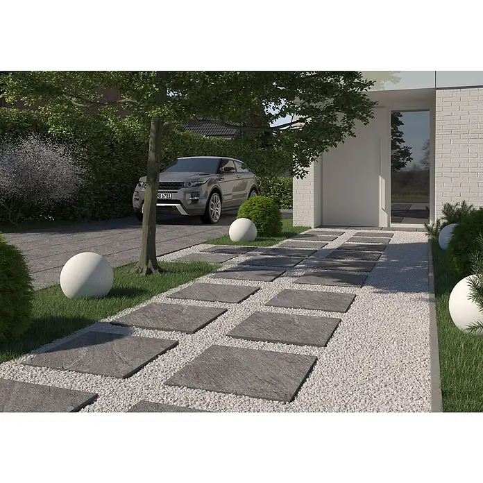 Terrassenfliese Piazza 2.0 - 59,7 x 59,7 x 2 cm, Anthrazit, Matt - Elegante Außenaufnahme mit einem modernen Gartenweg aus grauen Steinplatten, eingebettet in weißen Kies und üppiges Grün, führt zum Haus.