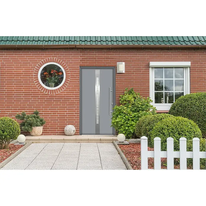 Splendoor Haustür Thermodoor PLUS SP20 - 110 x 210 cm, DIN Anschlag: Rechts, Verkehrsgrau - Fassade eines Hauses mit einer modernen grauen Haustür, einem runden Fenster und einem gepflasterten Gehweg, umgeben von üppigem Grün.