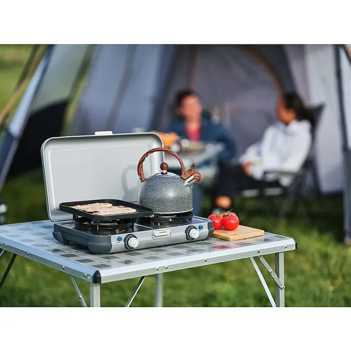 Campingaz Hornillo de gas 2 Grill & Go - 35 x 49 x 10 cm, 4 kW, Metal, Número de quemadores: 2 ud. - Cocina de camping con hornillo de gas, plancha y tetera sobre una mesa frente a una tienda. Perfecto para comidas al aire libre.