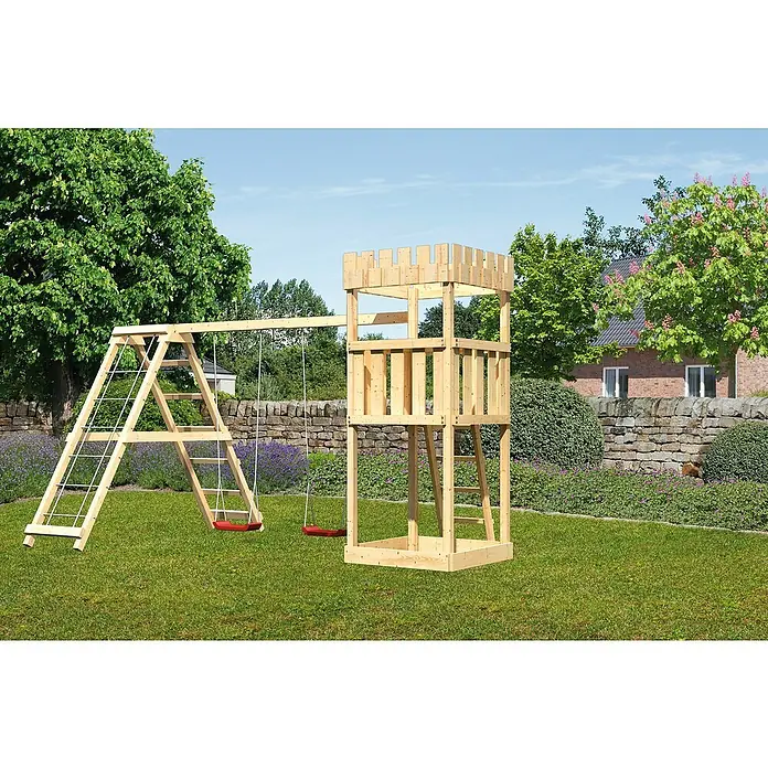 Karibu Kinderspielturm Ritterburg Löwenherz Set 5 - L x B x H: 264 x 415 x 259,5 cm, Natur - Ein hölzerner Spielturm mit Schaukel und Klettergerüst steht auf einer grünen Wiese, umgeben von Bäumen und Büschen.
