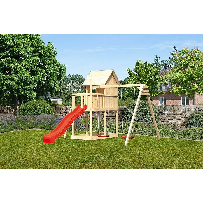 Karibu Kinderspielturm Frieda Set 28 - L x B x H: 264 x 347 x 291 cm, Natur - Ein hölzernes Spielhaus mit roter Rutsche und Schaukeln steht auf einem grünen Rasen vor Bäumen und einem Haus.