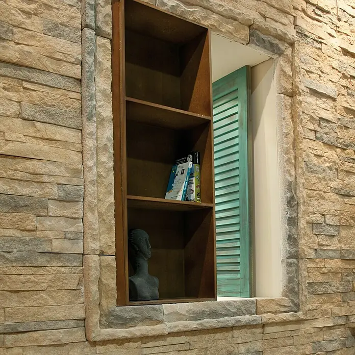 Revestimiento de piedra Salonica Canela - 60 x 30 x 2 cm - Estante con objetos decorativos integrado en una pared de piedra. La librería alberga libros y una escultura y está flanqueada por un marco de ventana.