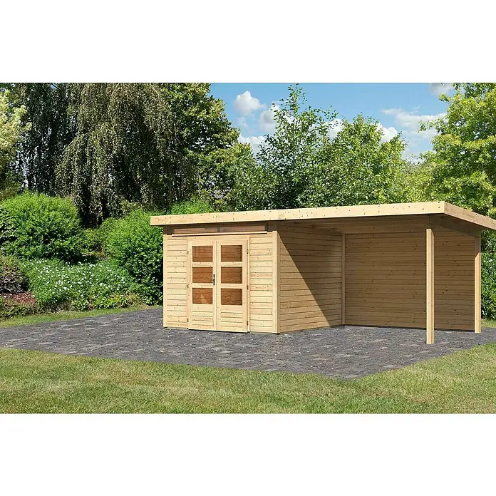 Karibu Gartenhaus Kandern 6 - Außenmaß inkl. Dachüberstand (B x T): 612 x 303 cm, Holz, Natur, Mit Seitendach und Rückwand - Ein Holzschuppen mit einem überdachten Bereich steht auf einem gepflasterten Untergrund, umgeben von Grün. Ideal für Gartengeräte und Lagerung.
