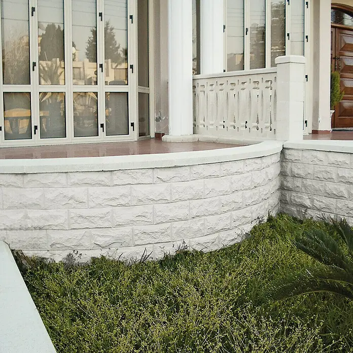 Revestimiento de piedra Teide - 14 x 28 cm, Blanco - Primer plano de una casa con una valla de piedra blanca que bordea un exuberante jardín. La fachada con ventanas refleja el paisaje circundante.