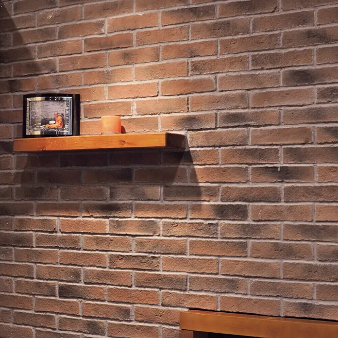 Revestimiento de piedra Mediterráneo Teja - 24 x 5,5 x 2,5 cm - Primer plano de una estantería de madera rústica con marco de fotos y vela sobre una pared de ladrillo. Ambiente cálido y acogedor en la habitación.