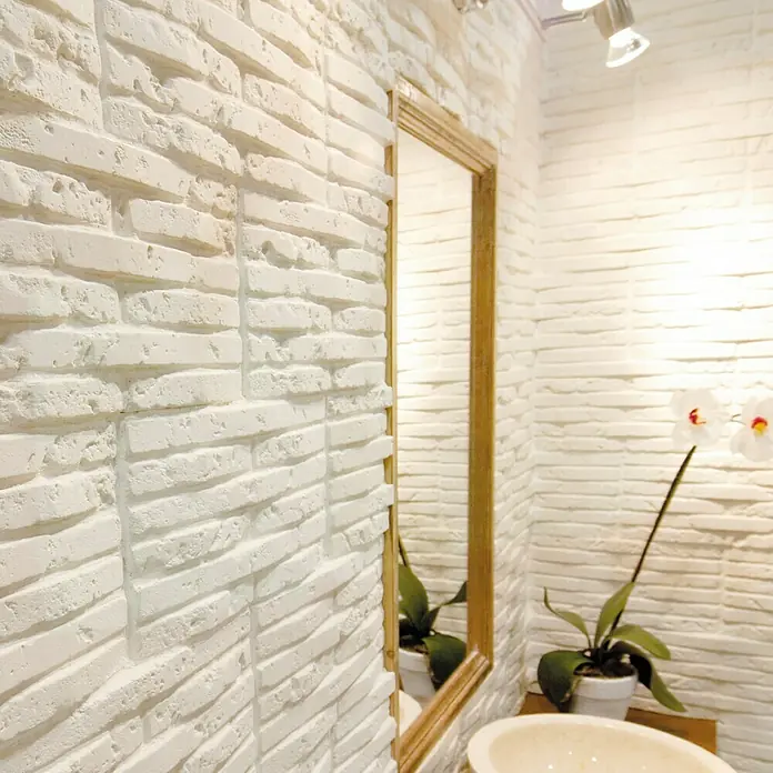 Revestimiento de piedra Listelo Hueso - 28 x 14 x 1 cm - Primer plano de un baño con paredes de ladrillo blanco, un espejo con marco de madera y una planta de orquídea blanca.