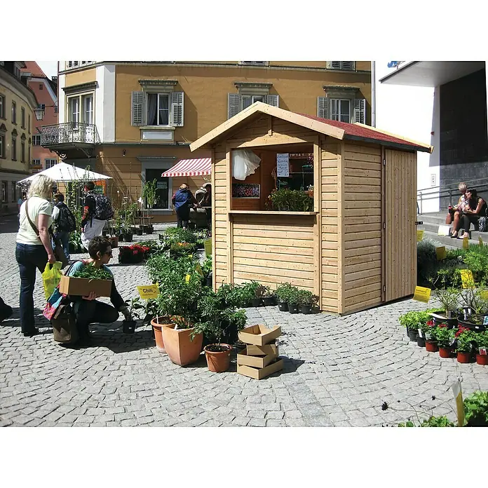 Karibu Marktstand - Außenmaß inkl. Dachüberstand (B x T): 290 x 216 cm, Holz, Natur - Außenansicht eines Verkaufsstandes aus Holz mit Pflanzen und Blumen, aufgestellt auf einem gepflasterten Platz.