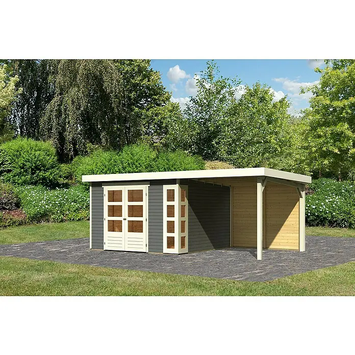 Karibu Gartenhaus Kerko 6Außenmaß inkl. Dachüberstand (B x T): 558 x 331 cm, Holz, Terragrau, Mit Seitendach und Rückwand - Ein graues Gartenhaus mit einem Carport-Anbau, beide mit cremefarbenen Akzenten, steht auf einem gepflasterten Bereich in einem üppigen Garten.
