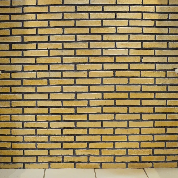 Revestimiento de piedra Mediterráneo Barro - 24 x 5,5 x 2,5 cm - Pared de ladrillos amarillos con juntas oscuras, que ofrece una textura rústica y cálida para fondos o representaciones arquitectónicas.