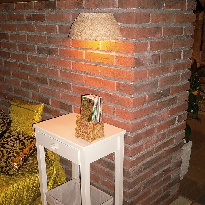 Esquina exterior Mediterráneo Teja - 24 x 12 x 5,5 cm - Una mesa auxiliar cuadrada y brillante hecha de madera está al lado de una pared de ladrillos con una lámpara y contiene algunos folletos en un recipiente.