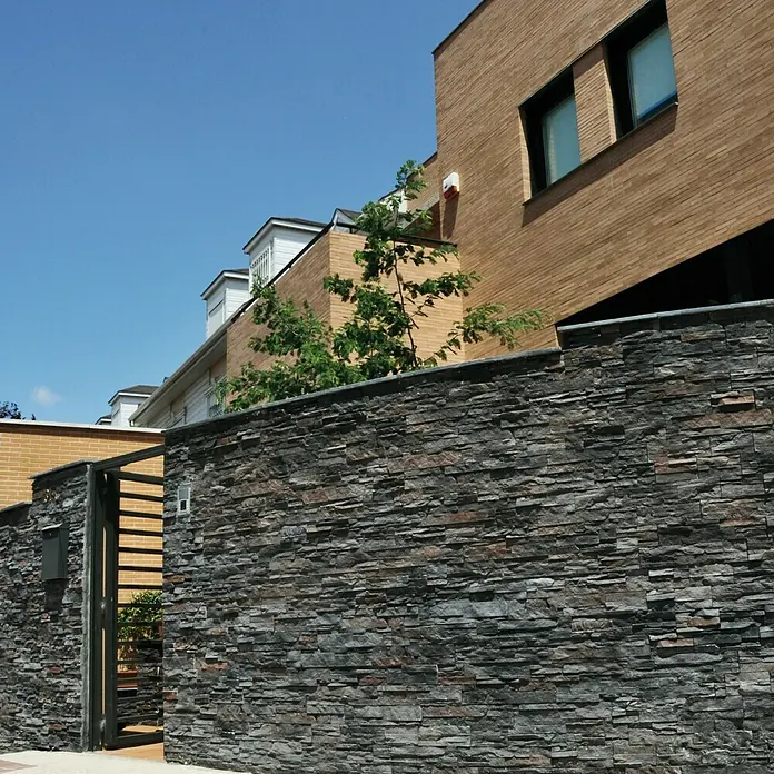 Revestimiento de piedra Petra Onix - Cantidad total: 4 pzs., Onix - Vista exterior de un muro de piedra con una puerta, que conduce a una casa moderna con arquitectura de ladrillo marrón.