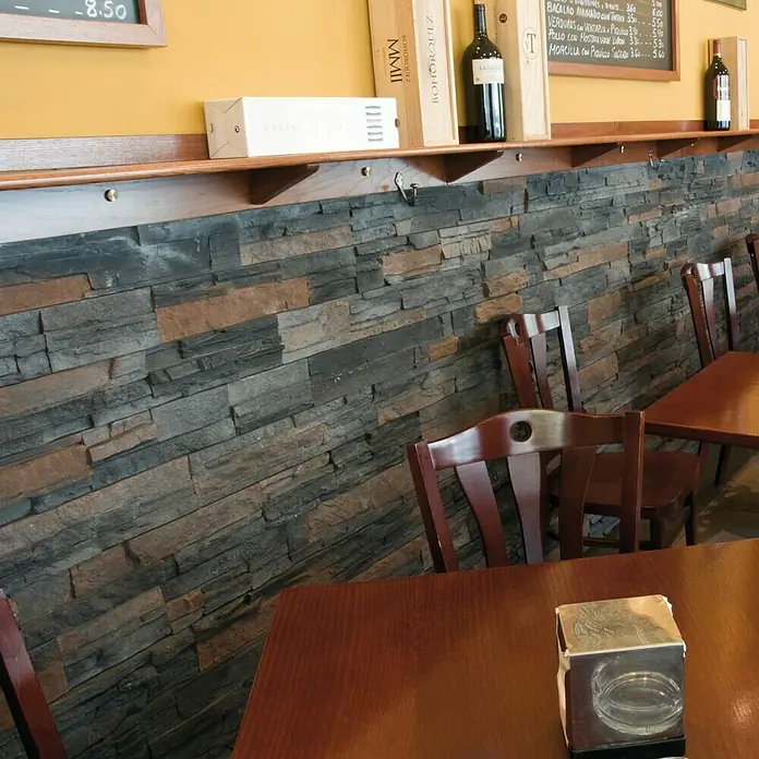 Revestimiento de piedra Petra Onix - Cantidad total: 4 pzs., Onix - Interior de un restaurante con mesas y sillas de madera frente a una pared de piedra con una estantería de madera que contiene botellas de vino.