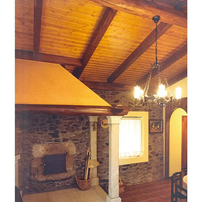 Panel de pared Frisolar Teñido - 240 cm x 95 mm x 10 mm - Interior rústico con chimenea de piedra y techo con vigas de madera, iluminado por una lámpara de araña clásica. Colores cálidos y diseño tradicional.